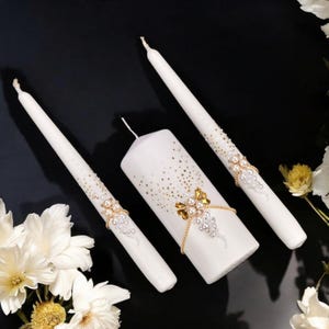 May include: Three white candles with decorative embellishments. The centerpiece is a pillar candle, flanked by two taper candles. All candles feature gold and pearl accents, with a scattering of gold flecks.