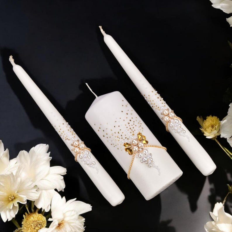 May include: Three white candles with decorative embellishments. The centerpiece is a pillar candle, flanked by two taper candles. All candles feature gold and pearl accents, with a scattering of gold flecks.