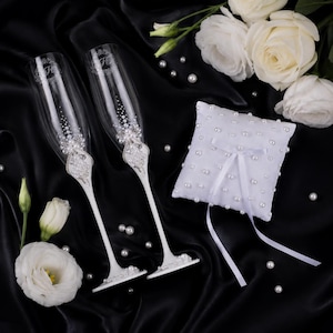May include: Two clear glass champagne flutes with white stems, decorated with white pearls and clear crystals, featuring an engraved monogram. A square white ring pillow adorned with scattered white pearls and a central white ribbon bow.