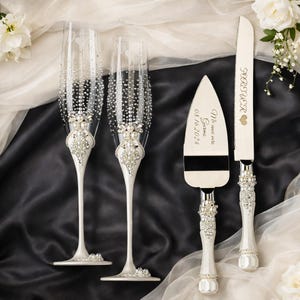 May include: Two clear champagne flutes with crystal and pearl embellishments on the stems and bowls, paired with a cake knife and server set featuring white handles with pearl accents. The server is engraved with Mr and Mrs Garma 08.16.2024.