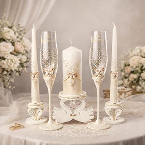 May include: Two clear glass champagne flutes and three off-white candles decorated with gold accents, pearls, and rhinestones. The center pillar candle rests on a white swan-shaped stand. The flutes feature gold text reading August 27, 2021.