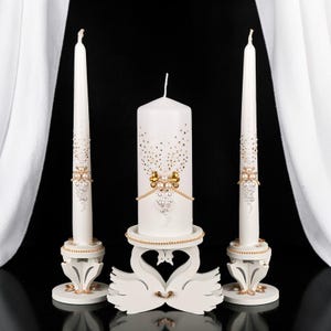 May include: A set of three white decorative candles. The tall, slender candles and the central pillar candle are adorned with gold and pearl accents. The pillar candle rests on a swan-shaped stand, adding an elegant touch. The candles are set against a black background.