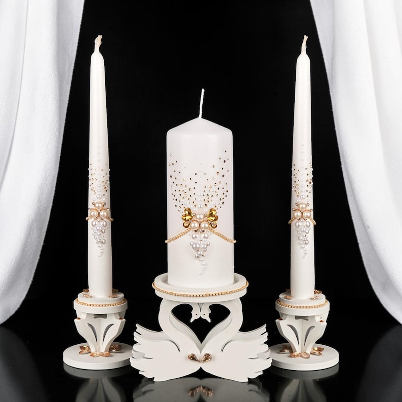 May include: A set of three white decorative candles. The tall, slender candles and the central pillar candle are adorned with gold and pearl accents. The pillar candle rests on a swan-shaped stand, adding an elegant touch. The candles are set against a black background.