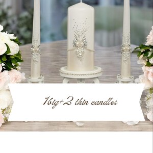 Op de afbeelding: Een set van drie gebroken witte kaarsen, bestaande uit &eacute;&eacute;n grote stompkaars en twee dunne dinerkaarsen, versierd met zilveren strass-steentjes en witte parels. De tekst "1 big + 2 thin candles" en "Where every detail makes history!" is zichtbaar.