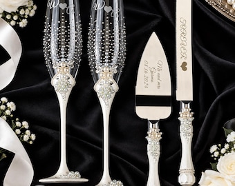 May include: Two clear glass champagne flutes with white stems, decorated with pearls and rhinestones, featuring engraved initials and hearts. A matching cake knife and server set with white handles, pearl accents, and engraved text including dates.