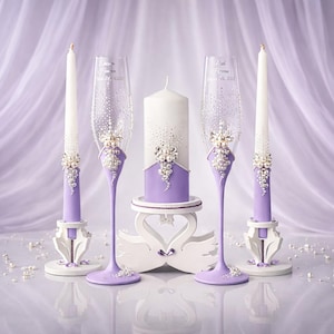 May include: A wedding unity candle set featuring two taper candles, a pillar candle, and two champagne flutes. The set is decorated with lavender paint, rhinestones, and pearls. The champagne flutes have text on them.