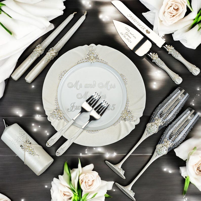 Op de afbeelding: Een huwelijksset met twee dinerkaarsen, een stompkaars, twee champagneglazen, een taartmes en een taartschep, versierd met strass-steentjes en parels. Een wit bord met de tekst Mr and Mrs ligt in het midden met twee zilveren vorken.