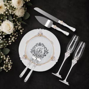 May include: A white ceramic plate with a gold rim and a central gray monogram letter K, two silver forks, a cake knife and server with white handles, and two clear glass champagne flutes, all featuring decorative silver and crystal embellishments.