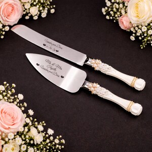 May include: A silver cake knife and server set with white handles adorned with pearls and gold accents. The knife and server are engraved with text and hearts. The set is displayed on a black surface, surrounded by floral arrangements.