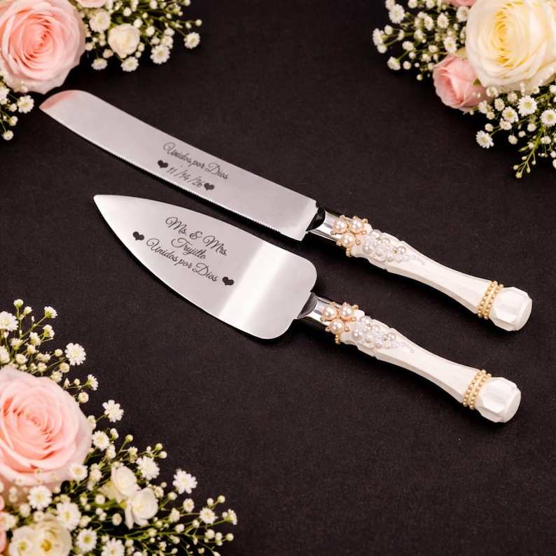 May include: A silver cake knife and server set with white handles adorned with pearls and gold accents. The knife and server are engraved with text and hearts. The set is displayed on a black surface, surrounded by floral arrangements.
