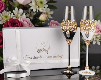 May include: Two champagne flutes, one black and one white, are adorned with gold and crystal embellishments. The flutes sit in front of a white gift box with the text "Two hearts - one destiny". A bouquet of flowers is in the background.