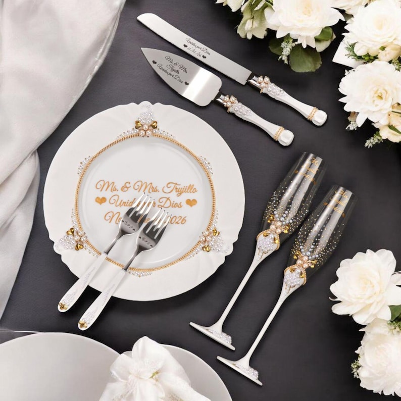May include: A wedding cake knife and server set, two champagne flutes, and a decorative plate with forks. The set is white and silver with gold accents and floral embellishments. The plate has gold text.