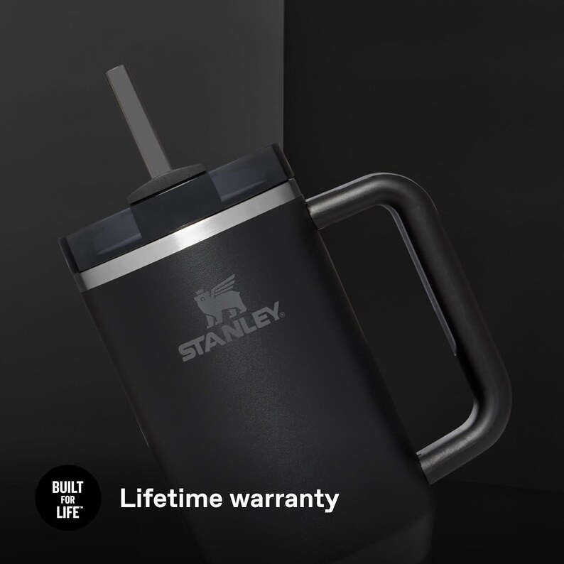 K&ouml;nnte beinhalten: Schwarzer Stanley-Becher mit Griff, Strohhalm und Deckel. Der Becher hat das Stanley-Logo und den Text "Lebenslange Garantie". Der Becher ist auf Langlebigkeit ausgelegt und ist f&uuml;r das Leben gebaut.