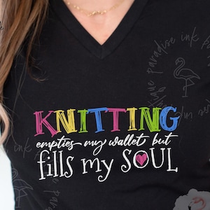 May include: Black V-neck t-shirt with the text "KNITTING empties my wallet but fills my SOUL" in colorful lettering. The word "KNITTING" is in rainbow colors. The shirt has short sleeves and a relaxed fit.