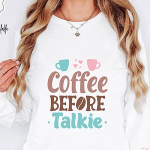 May include: White long-sleeved top with the phrase "Coffee Before Talkie" in brown, blue, and pink. The design includes coffee cup graphics and the text "More Colors Available" in a circular design.