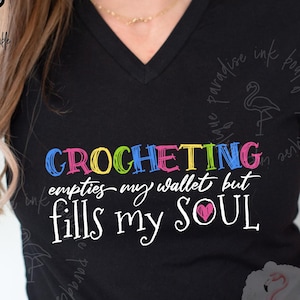 May include: Black V-neck t-shirt with the text "CROCHETING empties my wallet but fills my SOUL" in colorful letters. The shirt also has a pink heart graphic. "More Colors Available" is written on the top left.