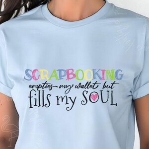 May include: Light blue t-shirt with the text "SCRAPBOOKING empties my wallet but fills my SOUL" in colorful lettering. The shirt has short sleeves and a round neckline. A light blue circle with the words "Light Blue" is in the upper left corner.