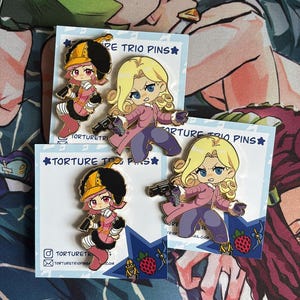 Puede incluir: Dos juegos de pines de esmalte con personajes estilo anime sobre tarjetas blancas con la etiqueta TORTURE TRIO PINS. Un pin muestra a un personaje con traje rosa y sombrero amarillo, el otro a un personaje rubio con abrigo rosa sosteniendo una pistola negra.