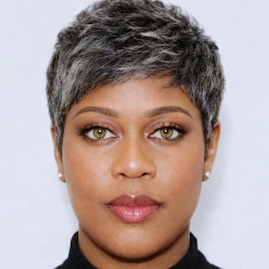 May include: A person with short, grey and white hair and green eyes, wearing a black polo neck. The image is a portrait against a plain white background.
