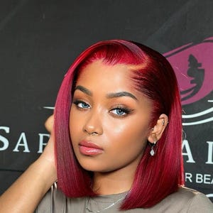 May include: A woman with bright blue eyes and a bob hairstyle dyed a vibrant red color. She is wearing a silver necklace and has multiple piercings. The background is dark with white text that is partially visible.