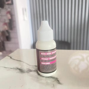 Puede incluir: Una pequeña botella blanca con tapa blanca. La etiqueta presenta texto en rosa y blanco, incluyendo las palabras "Water Based Copolymer" y "Specially Formulated". La botella está sobre una superficie clara.