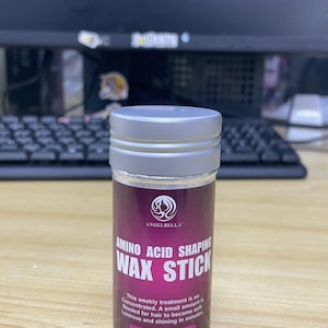 May include: A cylindrical hair styling product labeled "ANGELBELLA AMINO ACID SHAPING WAX STICK." The container is silver and burgundy with white text. It states "STRONG HOLD" and contains 75g or 2.7 fl oz of product. The product promises soft, lustrous, and shining hair.