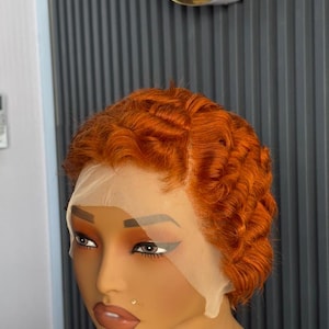 Könnte beinhalten: Eine kurze, orangefarbene Perücke mit Spitzenfront wird auf einem Schaufensterkopf gezeigt. Die Perücke hat einen welligen, strukturierten Stil. Die Schaufensterpuppe hat Make-up, einschließlich Lidschatten und Lippenstift. Der Hintergrund ist eine graue Wand.