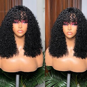 May include: Two black curly wigs with bangs are displayed on mannequin heads. The wigs have a natural-looking texture and are styled with a fringe. The mannequins are set against a backdrop of wooden panels and greenery.