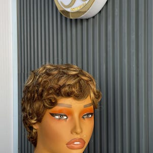 May include: A mannequin head with a short, curly brown wig with blonde highlights. The mannequin has full makeup, including orange eyeshadow and dark eyeliner. The background is a grey, vertically-lined wall.