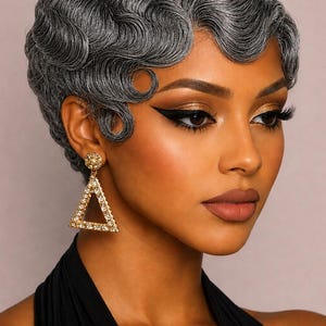 May include: A woman with a short, grey, wavy hairstyle and gold triangle earrings. She wears dramatic eye makeup and a black top. The earrings are encrusted with small, clear stones.