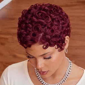 May include: A close-up of a person with short, curly, burgundy-colored hair. The hair is styled in tight, defined curls. The person is wearing a silver chain necklace and hoop earrings. The person is wearing a white top.