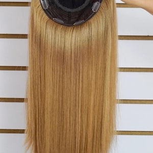 May include: A blonde hair topper with a black base and multiple clips. The hair is long, straight, and a warm shade of blonde. The topper is held up against a white background with horizontal wooden slats.