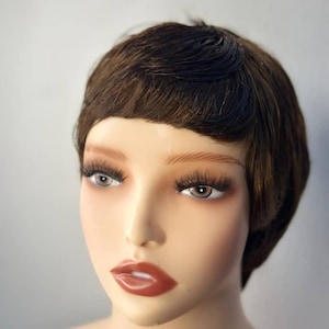 May include: A mannequin head with a short, brown wig styled in a classic pixie cut. The mannequin has full eyebrows, long eyelashes, and glossy red lipstick. The skin tone is fair, and the background is a solid gray.