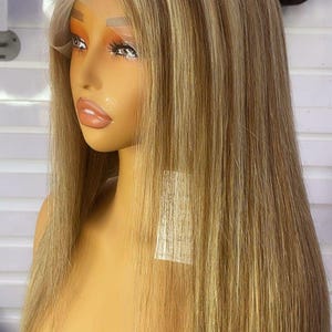 Pode incluir: Uma cabeça de manequim exibindo uma peruca longa e lisa. A peruca apresenta uma mistura de loiro e castanho claro, com uma frente de renda. O cabelo é penteado com uma risca ao meio e cai até os ombros.