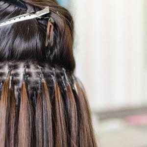May include: Close-up of hair extensions attached to natural hair. The extensions are a blend of brown and auburn shades, secured with small, dark attachments. A silver hair clip is visible at the top, holding back some of the hair.