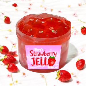 May include: A clear plastic container filled with red strawberry-flavored jelly with small strawberry pieces. The container has a pink label that reads "Skye's Strawberry JELLO Do not eat."