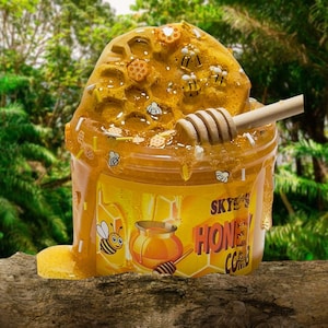 May include: A yellow honey comb slime in a clear container with a wooden honey dipper. The container has a label that says "Skye's Honey Comb".