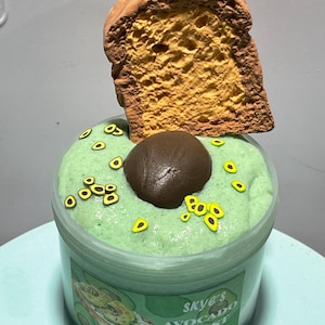 May include: A clear plastic container filled with green slime, topped with a brown chocolate avocado and a slice of brown bread. The container has a label that reads "Skye's Avocado Toast" and "Do not eat".