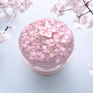 May include: A jar of pink slime decorated with white and gold sprinkles and small white flowers. A large white and pink flower is centered on top. The jar is set against a light blue background with cherry blossom branches.