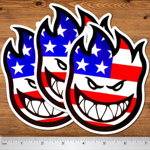 May include: Three stickers with a flame design, featuring the American flag's stars and stripes. The flames have a black outline and a grinning face with sharp teeth. The stickers are on a wooden surface.