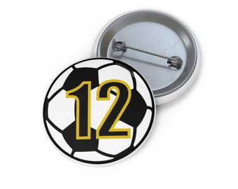 May include: A round button with a soccer ball design. The soccer ball is black and white with the number 12 in gold. A second button is visible in the background.