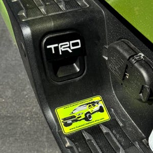 May include: A green Toyota Tacoma truck with a black "TRD" emblem on the tailgate. A yellow sticker with a black and white illustration of a truck and the text "www.GuacosCreations.com" is on the tailgate.