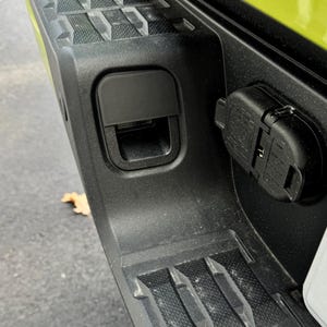 May include: A close-up of a black plastic truck bed with a black plastic cover over a trailer hitch. The cover has a small opening for a trailer hitch pin. The truck bed has a textured surface.