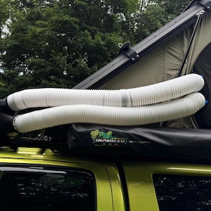 May include: A yellow vehicle with a rooftop tent and a black storage box. Two white, flexible tubes are attached to the storage box and extend over the tent. The tent is olive green and black. The image is taken outdoors with trees in the background.