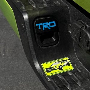 May include: A green Toyota Tacoma truck with a black plastic bumper. The bumper has a black plastic latch with a blue "TRO" emblem. A yellow sticker with a black and white image of a truck and the text "www.GuscasCreations.com" and "www.GuscasCreations.etsy.com" is on the bumper.