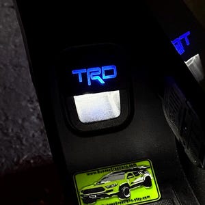 May include: A black car door with a blue illuminated "TRD" emblem and a green sticker with the text "www.SunsetCreations.com" and a picture of a green truck.
