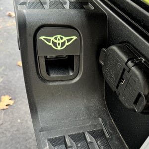 May include: A black truck tailgate with a green Yoda logo on a black plastic cover. The cover has a small rectangular opening with a black plastic latch. A black plastic connector is visible on the right side of the image.