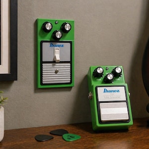 May include: Two green Ibanez guitar effects pedals with black knobs and silver accents. One pedal is mounted on a wall, the other is on a wooden surface. Guitar picks and a framed picture are also visible.