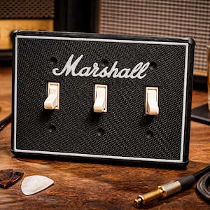 May include: Black Marshall-branded switch plate with three cream-colored toggle switches. The plate has a white border and is set against a wooden surface. A glass of amber liquid, headphones, and a guitar cable are also visible.