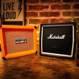 May include: Two miniature guitar amplifiers, one orange with "Orange" text and the other black with "Marshall" text. The amps sit on a wooden surface with a brick background and a "Live Loud" sign.